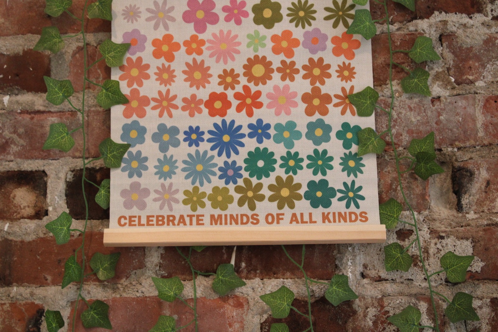 A print reading Celebrate Minds Of All Kinds, covered in rows of coloured flowers, hanging on an exposed brick wall with ivy trailing across it.