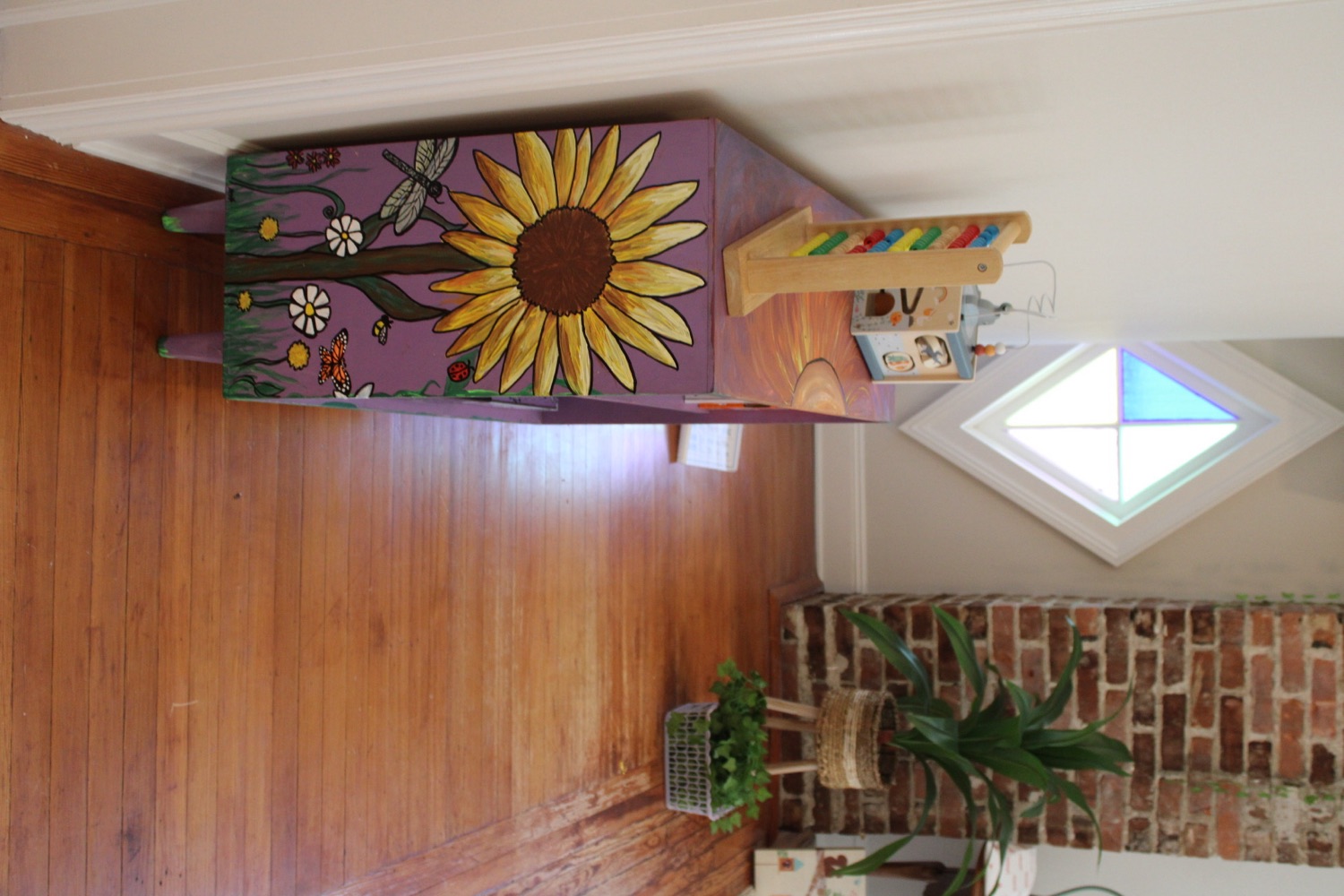 The hallway outside the Grow Space room, with a hand painted purple dresser, a stained glass window and an exposed brick wall.
