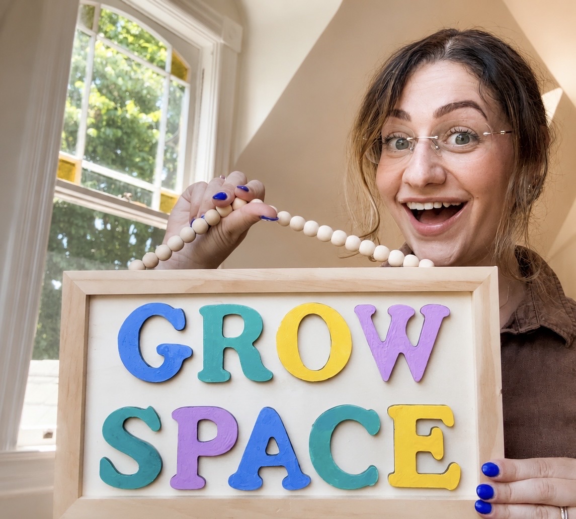 Linda smiling as she holds up the hand painted Grow Space sign, with wooden letters in blue, teal, gold and lilac.
